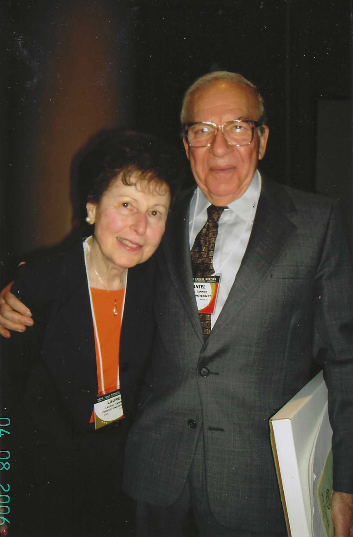 Two elderly individuals pose together, smiling warmly at a gathering, dressed in formal attire. The woman wears pearls and an orange blouse,
