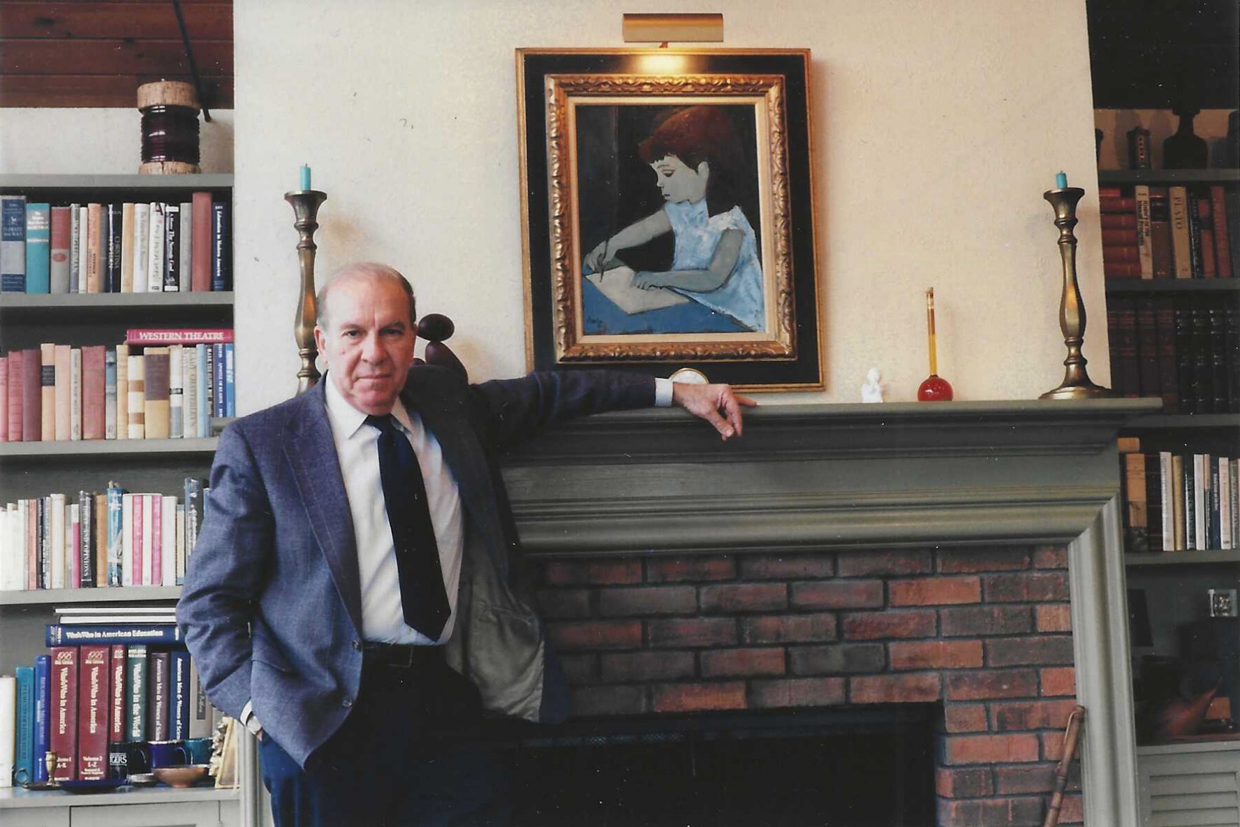A man in a suit leans against a brick fireplace, with a portrait of a girl in a blue dress on the wall behind him